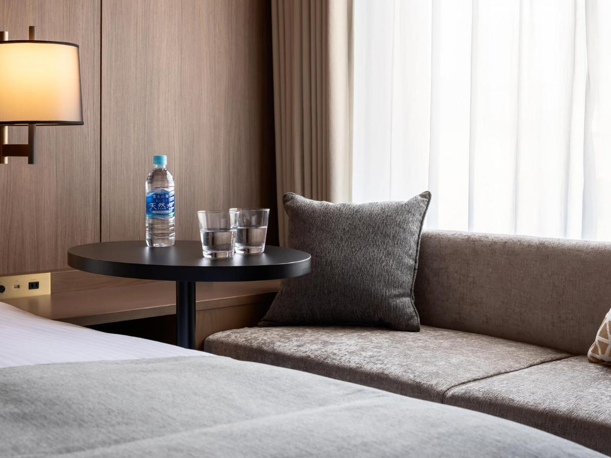 Mitsui Garden Hotel Roppongi Tokyo Premier Экстерьер фото The photo depicts a cozy hotel room setting. On the left, there is a bedside table with a lamp. Against the wall, there is a comfortable-looking couch with a decorative pillow. In front of the couch, there's a small round table that holds a bottle of
