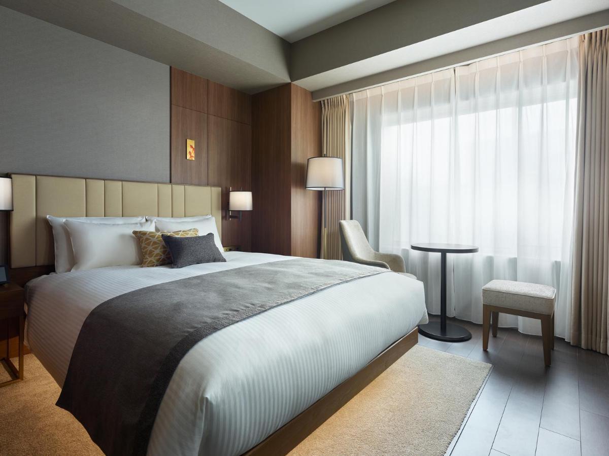 Mitsui Garden Hotel Roppongi Tokyo Premier Экстерьер фото The photo shows a modern hotel room featuring a comfortable bed with white linens and a decorative throw pillow. The room has a neutral color palette with wooden accents on the walls. There are two bedside lamps and a chair positioned next to a small