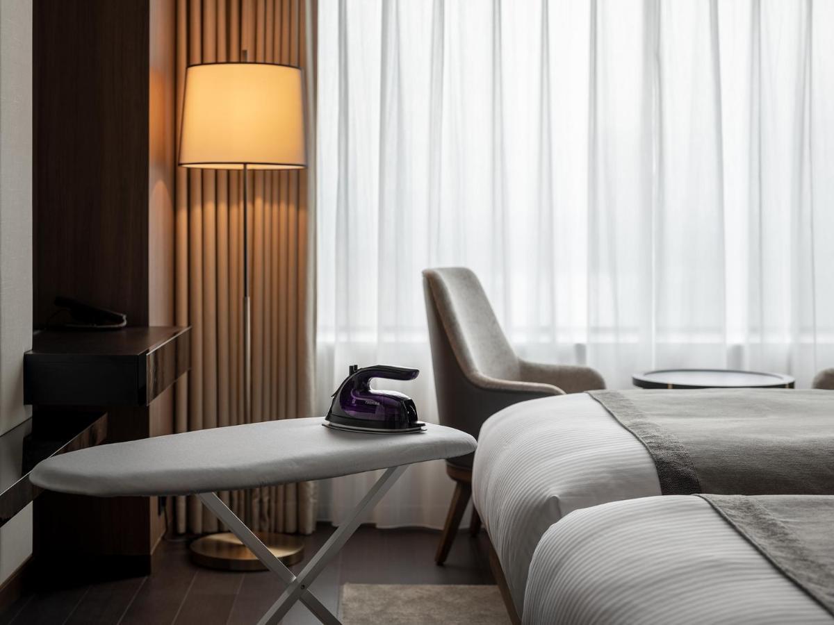 Mitsui Garden Hotel Roppongi Tokyo Premier Экстерьер фото The photo shows a modern hotel room featuring two beds with white bedding. There is a light-colored armchair next to a small side table. On the table, there is an iron, and beside it, a lamp with a warm glow is illuminating the space. The large windo