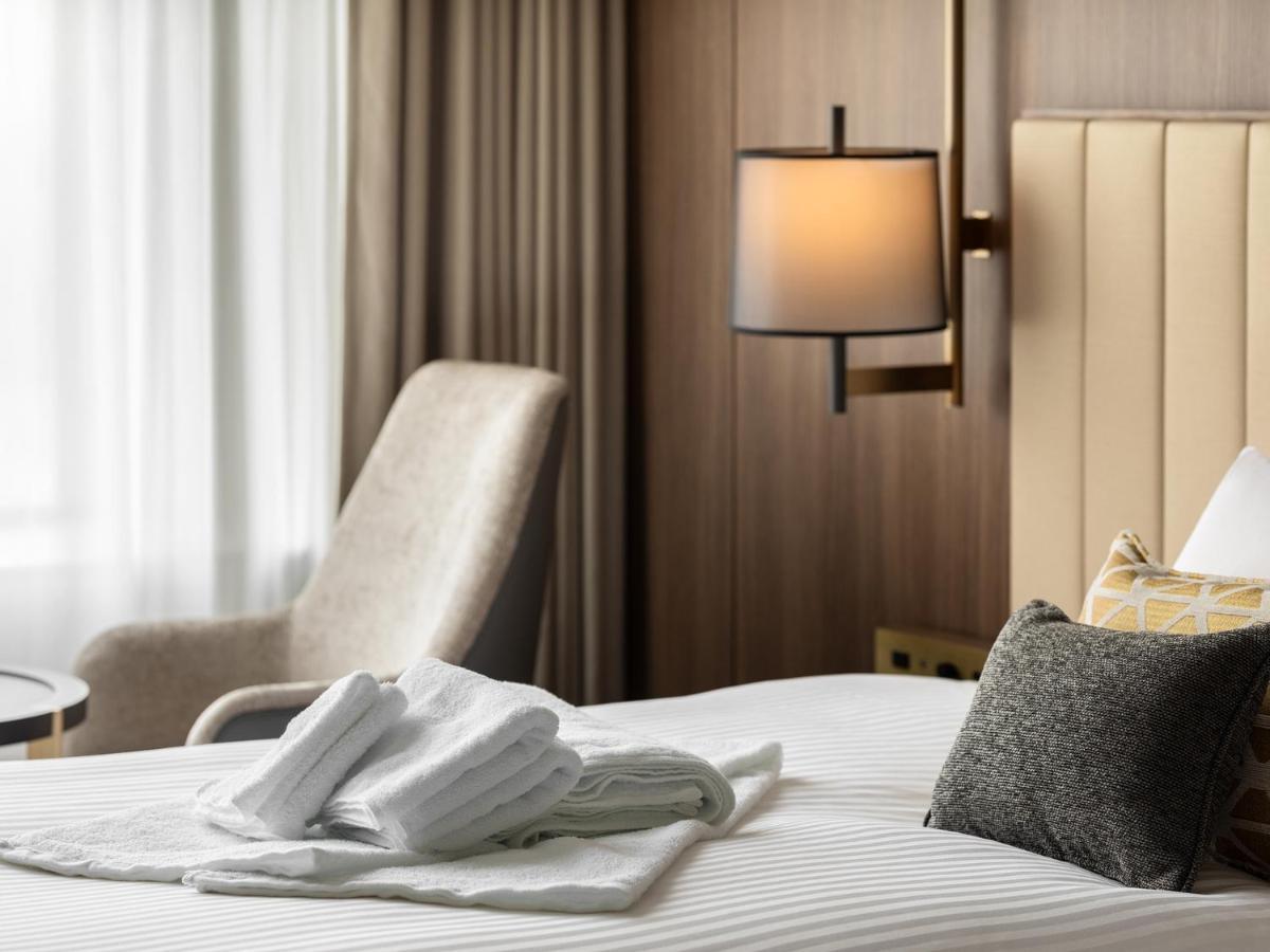 Mitsui Garden Hotel Roppongi Tokyo Premier Экстерьер фото The image shows a stylish hotel room. In the foreground, there is a neatly made bed with white linens, featuring a stack of white towels arranged in a decorative manner. To the side, there is a gray upholstered armchair and a small table. The backgro