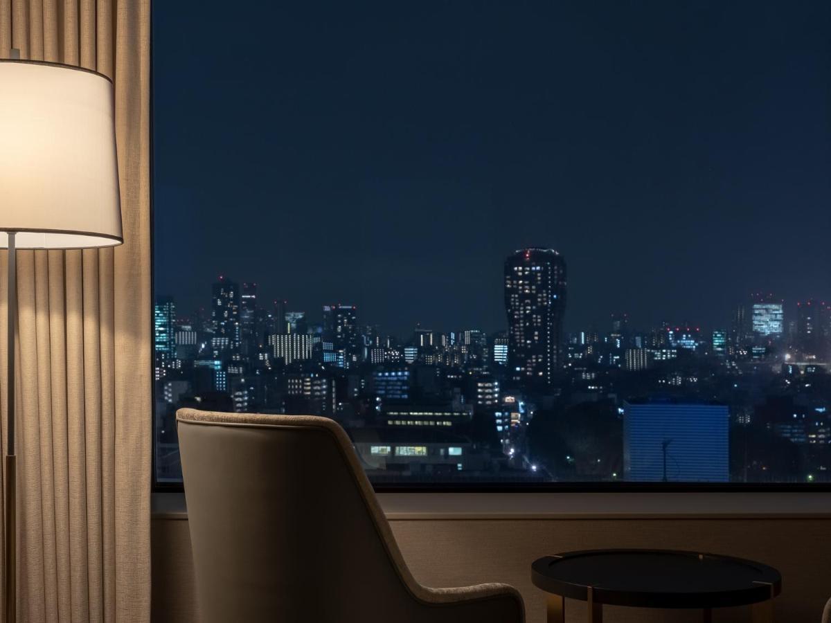 Mitsui Garden Hotel Roppongi Tokyo Premier Экстерьер фото The photo depicts a cozy hotel room setting at night. In the foreground, there's a stylish armchair next to a small round table. A standing lamp provides soft lighting, creating a warm ambiance. In the background, large windows reveal a city skyline 