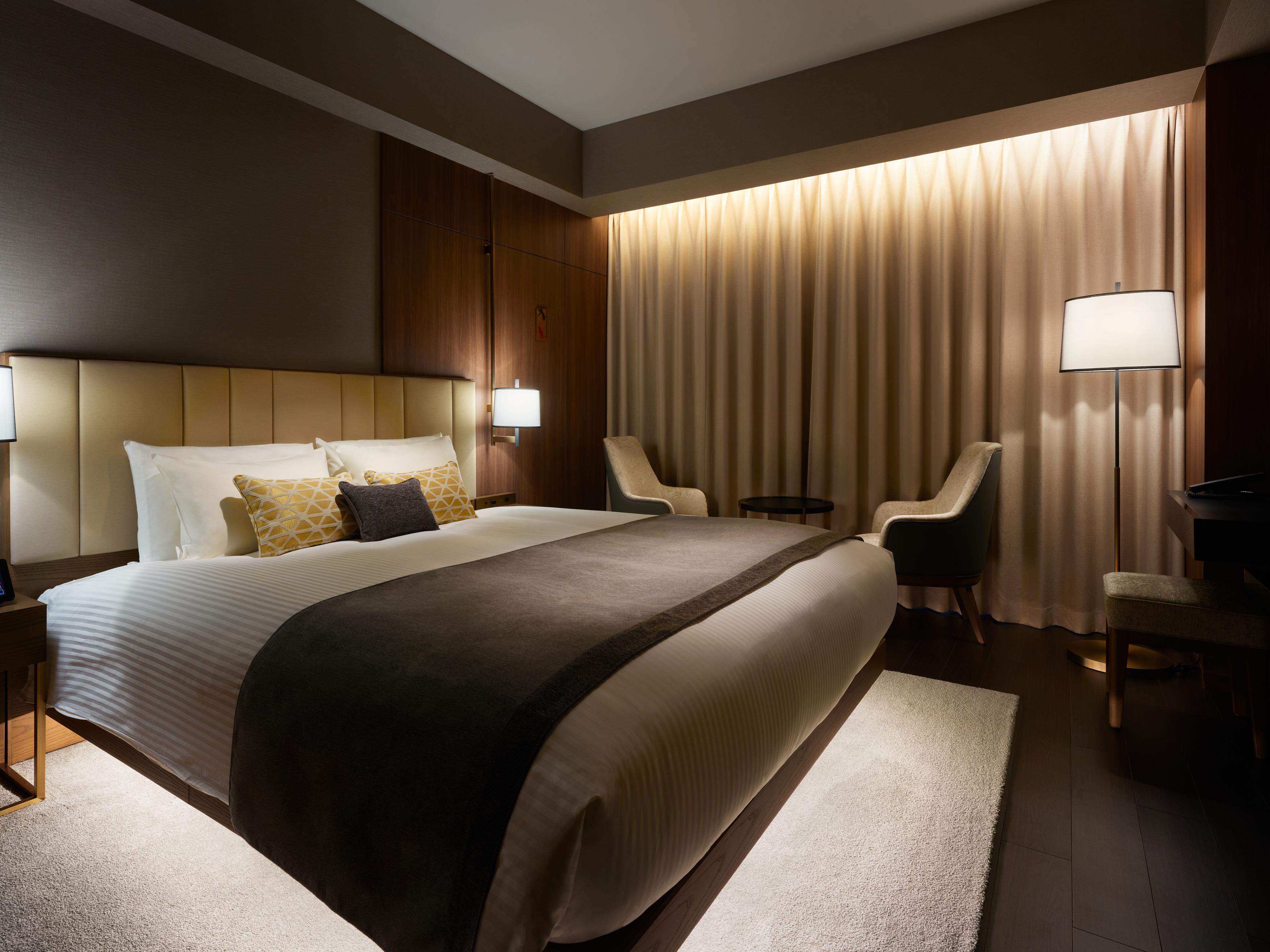 Mitsui Garden Hotel Roppongi Tokyo Premier Экстерьер фото The photo depicts a modern hotel bedroom. It features a large bed with white linens and a dark gray throw blanket, accented by decorative pillows. The room has a warm ambiance created by soft lighting, including two bedside lamps and a lighted area b