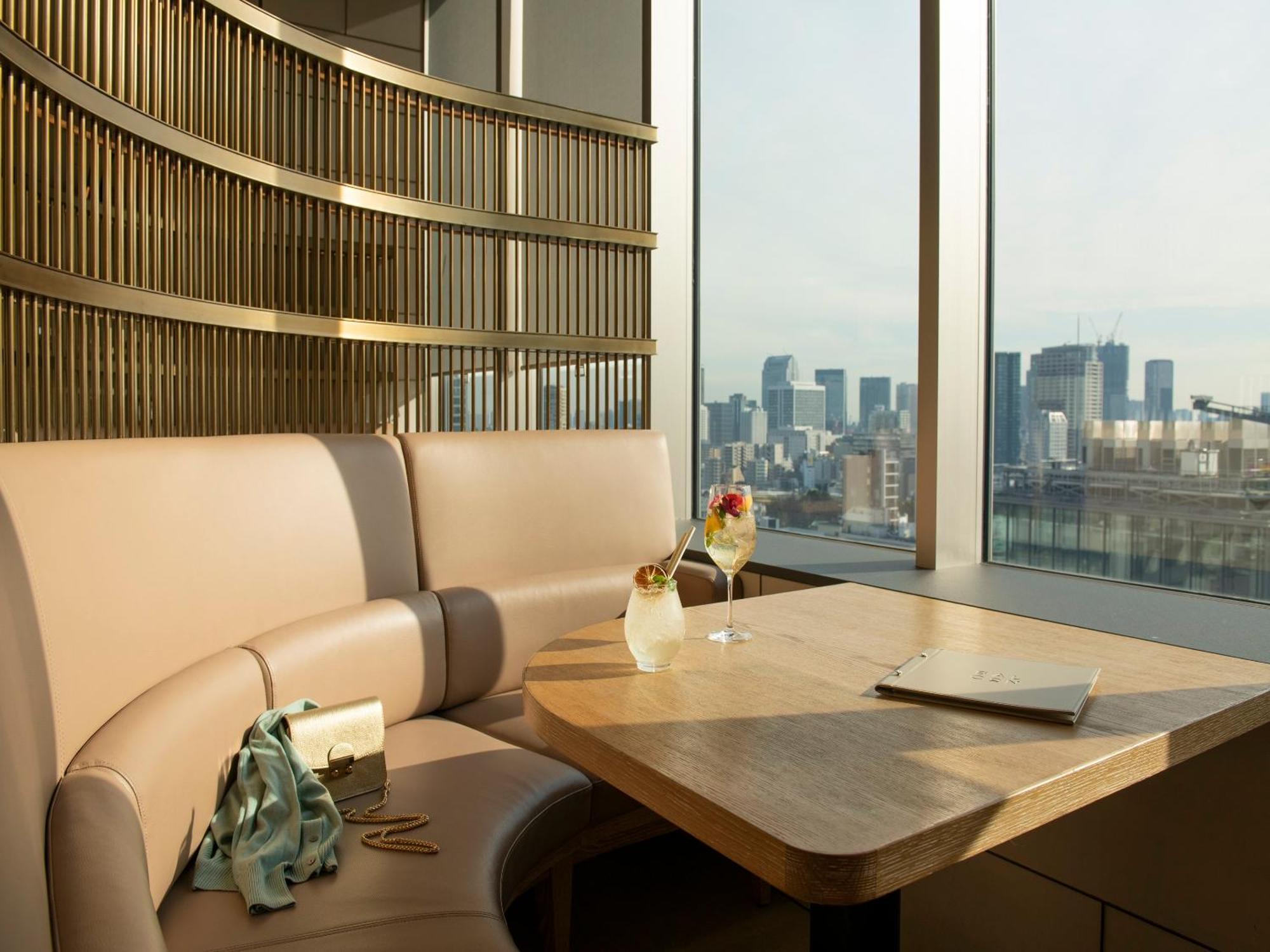 Mitsui Garden Hotel Roppongi Tokyo Premier Экстерьер фото The photo depicts a cozy, modern seating area with a curved booth that features soft, pale upholstery. A wooden table is positioned in front of the booth, and on the table, there are two elegant drinks garnished with fruit. The setting has large wind