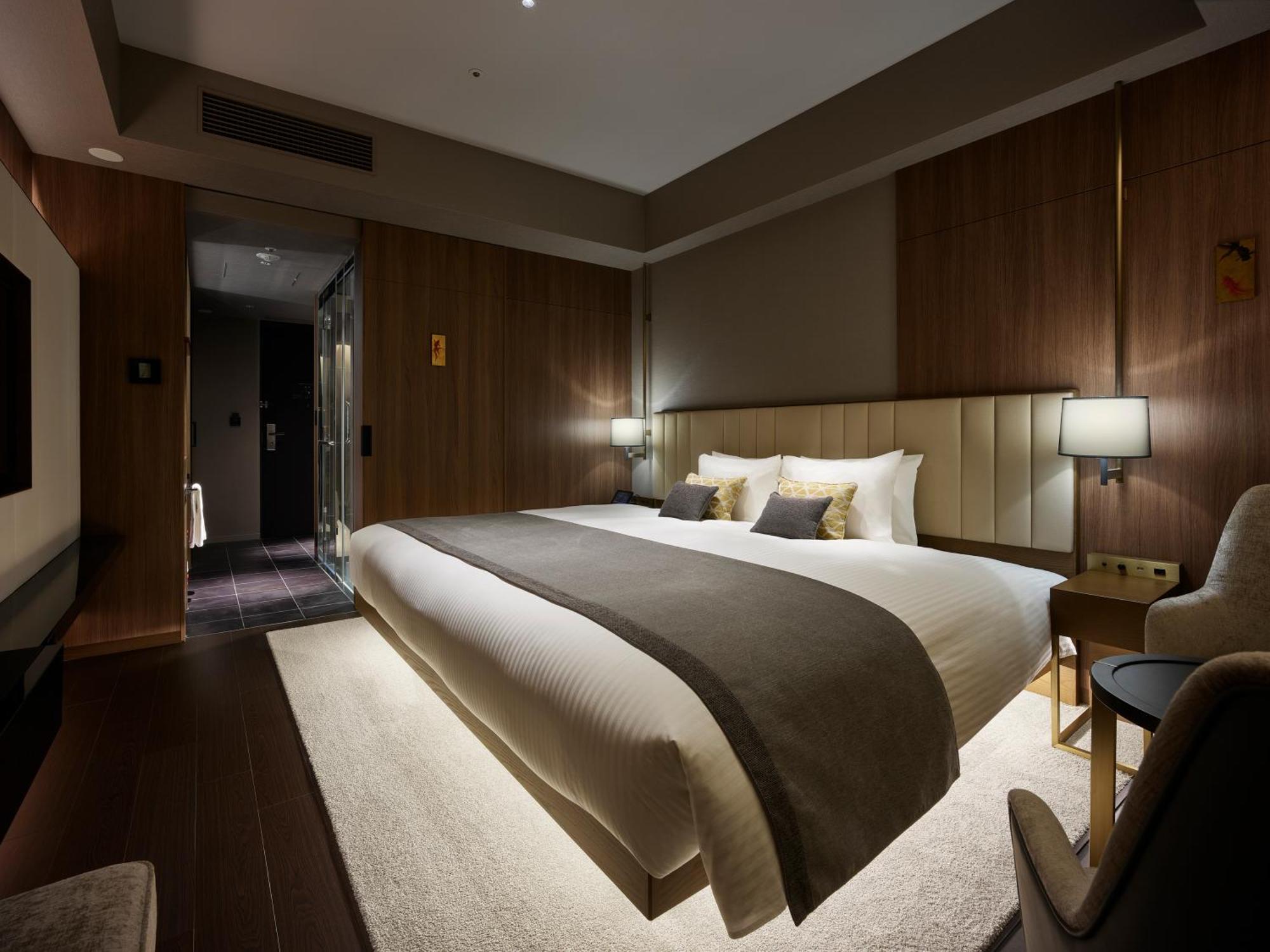 Mitsui Garden Hotel Roppongi Tokyo Premier Экстерьер фото The photo shows a modern hotel room featuring a large bed with a neutral-colored duvet and a dark throw blanket. The headboard is upholstered, and there are two bedside lamps on either side of the bed. The walls have a warm wood paneling, contributin