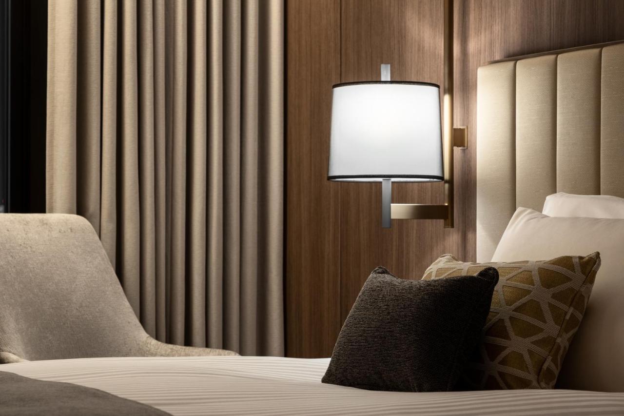 Mitsui Garden Hotel Roppongi Tokyo Premier Экстерьер фото The photo shows a cozy hotel bedroom setting. There is a neatly made bed with white linens and a few decorative pillows, including a dark one and another with a geometric pattern. To the side, there is a soft armchair and a modern wall lamp mounted a